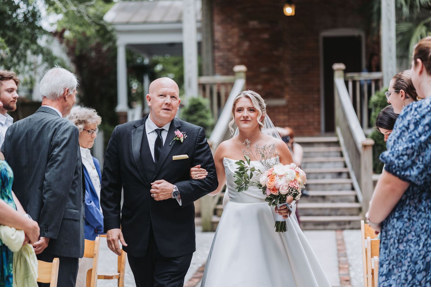 Father walks smiling bride down outdoor aisle as guests watch during wedding ceremony.