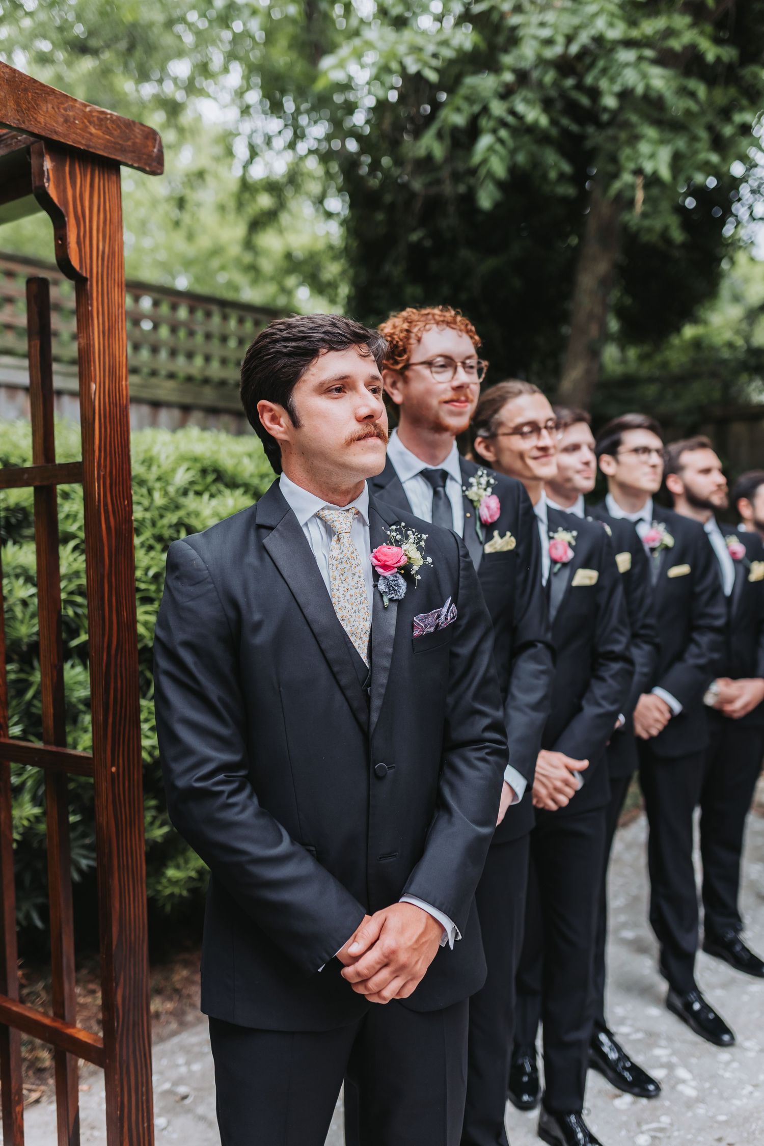 Groom and groomsmen in dark suits lined up outdoors awaiting bride at garden wedding ceremony.