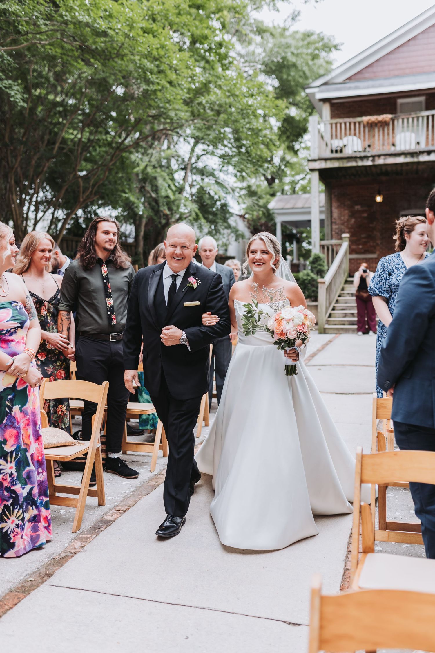 A smiling bride in a white ball gown walks down the outdoor aisle with her father.