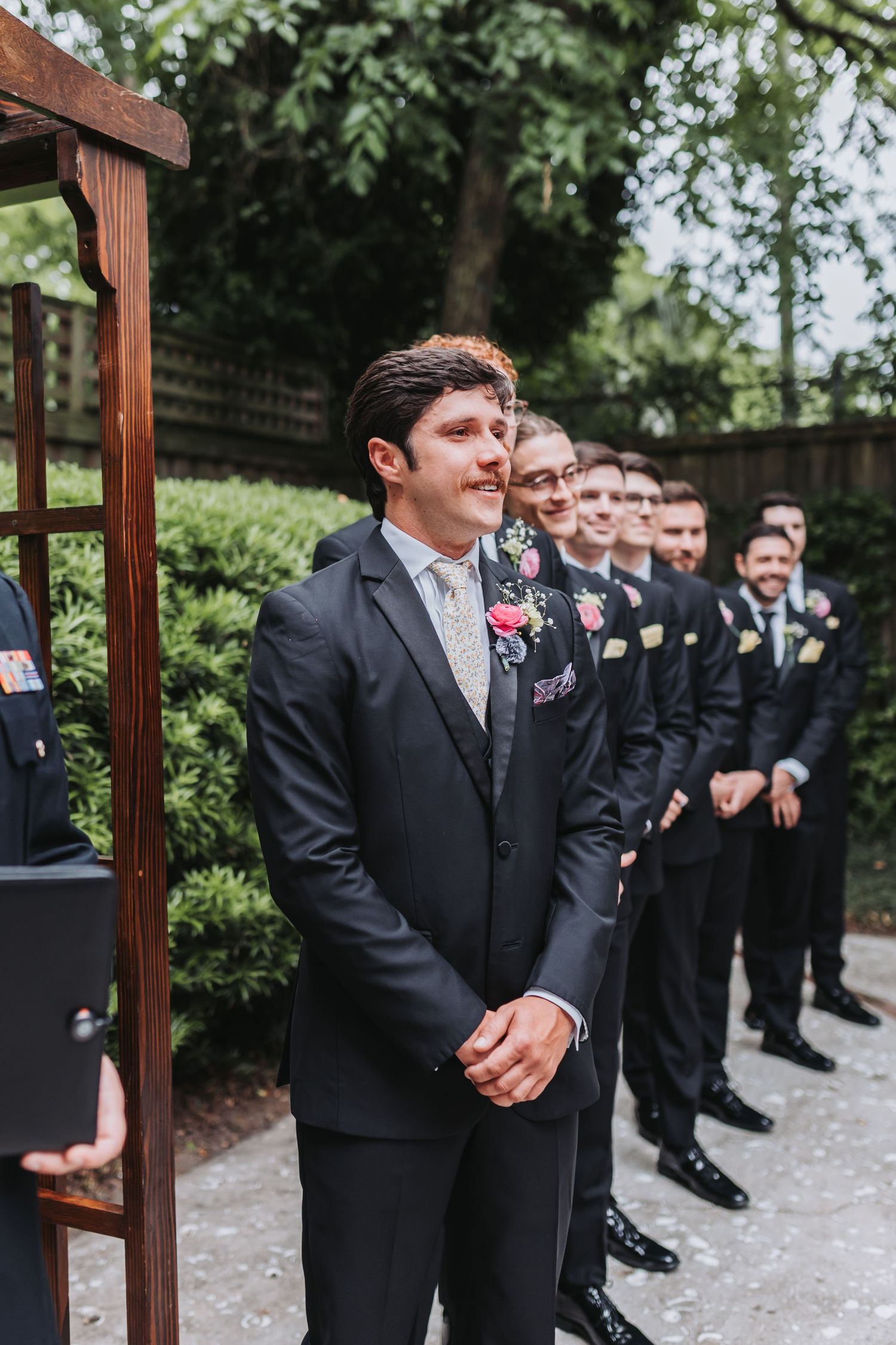 Groom smiles warmly at altar with groomsmen lined up during outdoor wedding ceremony.
