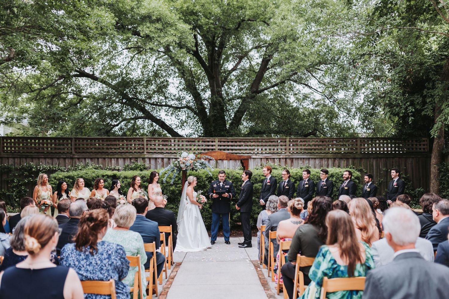 A wedding ceremony takes place outdoors under a large tree with guests seated on wooden chairs.