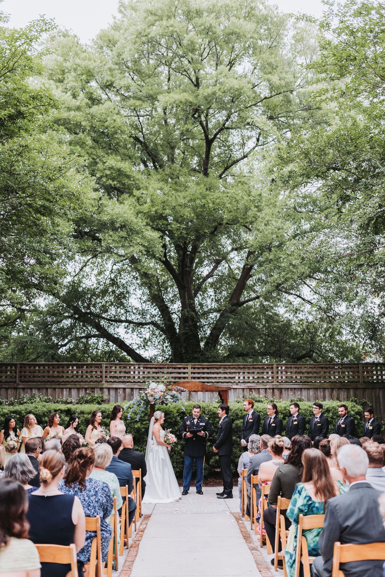 Outdoor wedding ceremony with guests seated along an aisle, couple at altar beneath a large green tree.