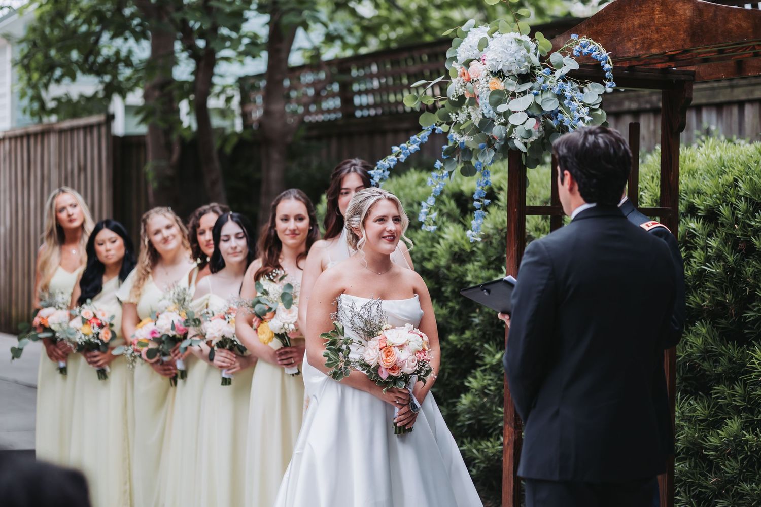Bride stands with bridesmaids in yellow dresses holding colorful bouquets during outdoor wedding ceremony.