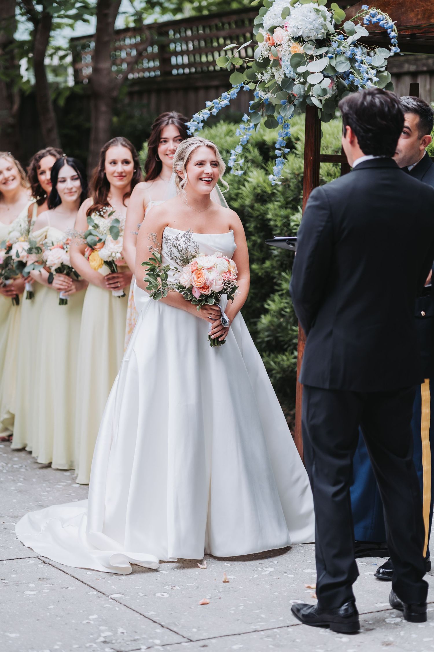 Smiling bride in strapless white gown stands at altar with bridesmaids in yellow dresses holding bouquets.
