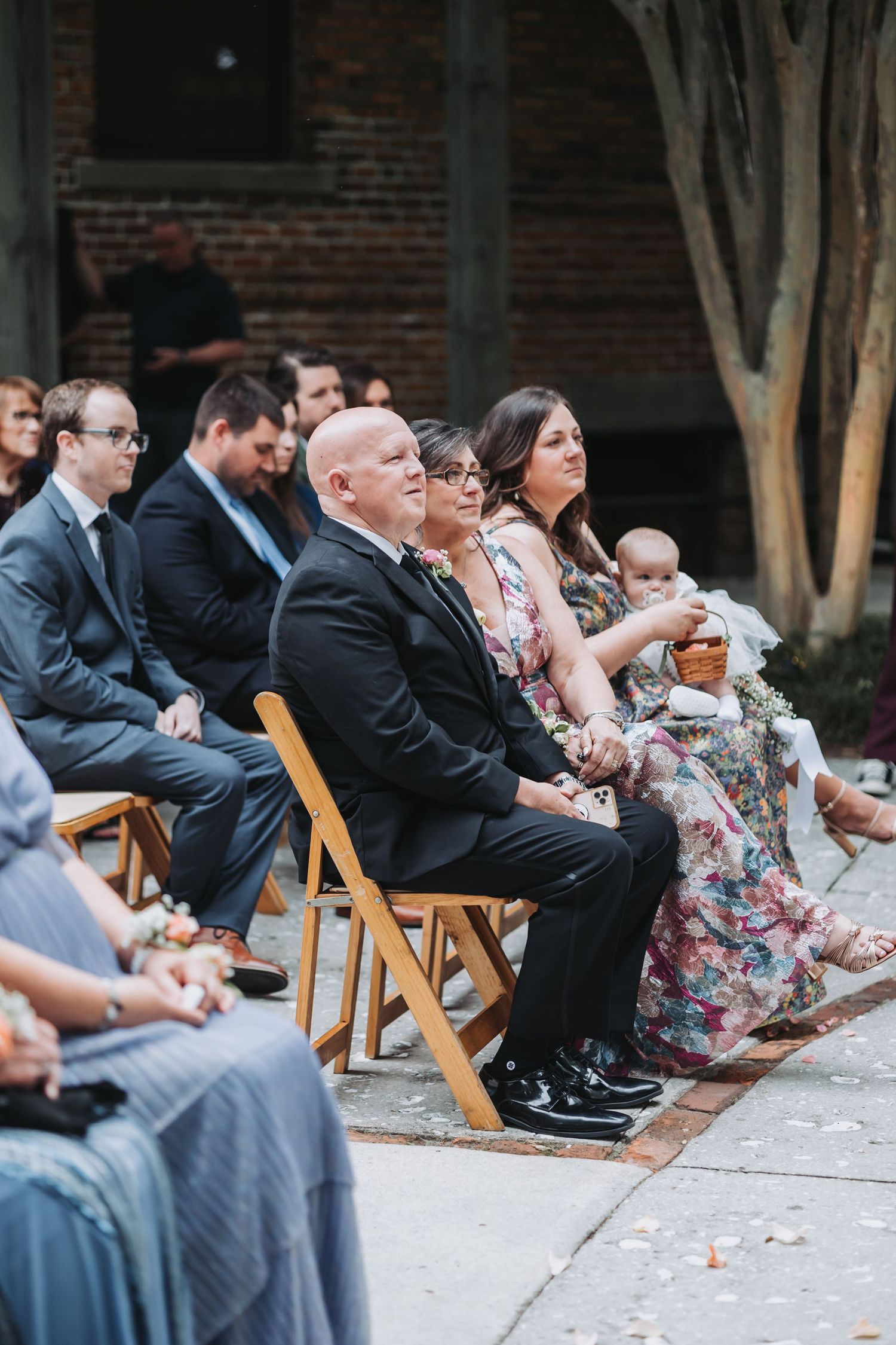 Wedding guests seated in wooden chairs watch outdoor ceremony including woman holding baby.