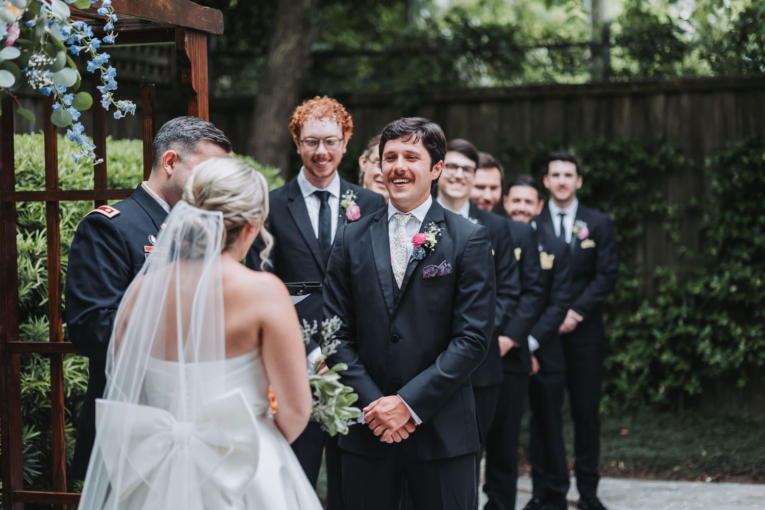 Groom smiles at bride approaching altar with groomsmen standing in line at outdoor ceremony.