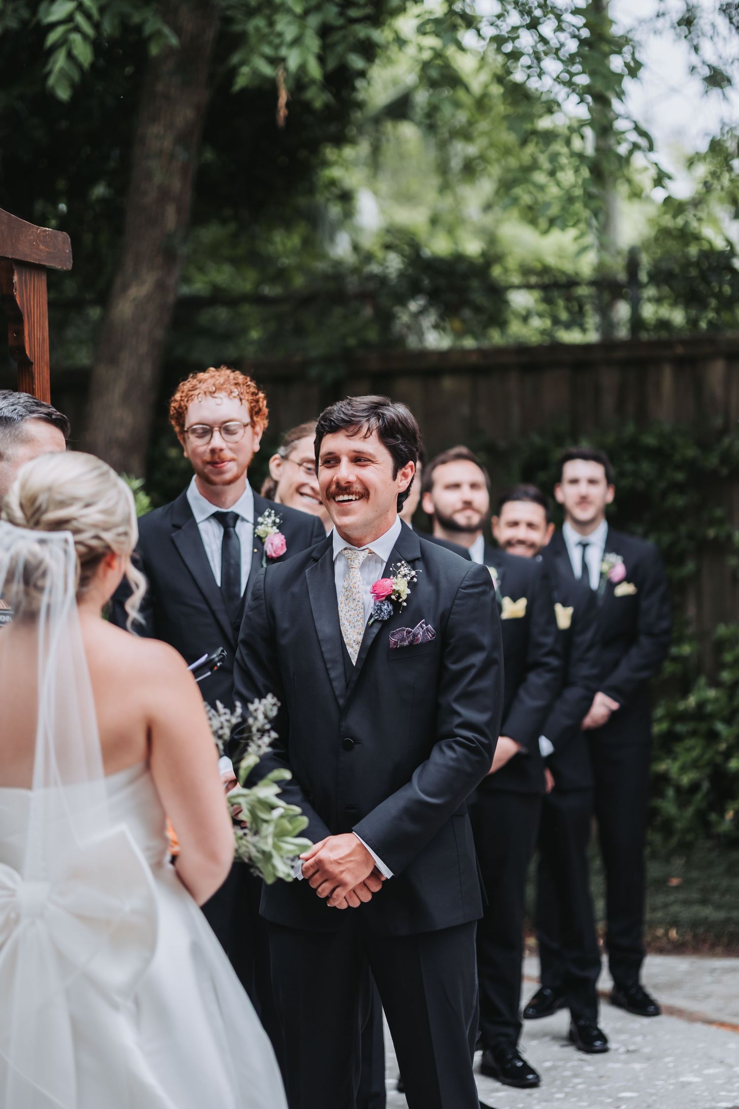 Groom smiles at bride approaching altar with groomsmen lined up outdoors.