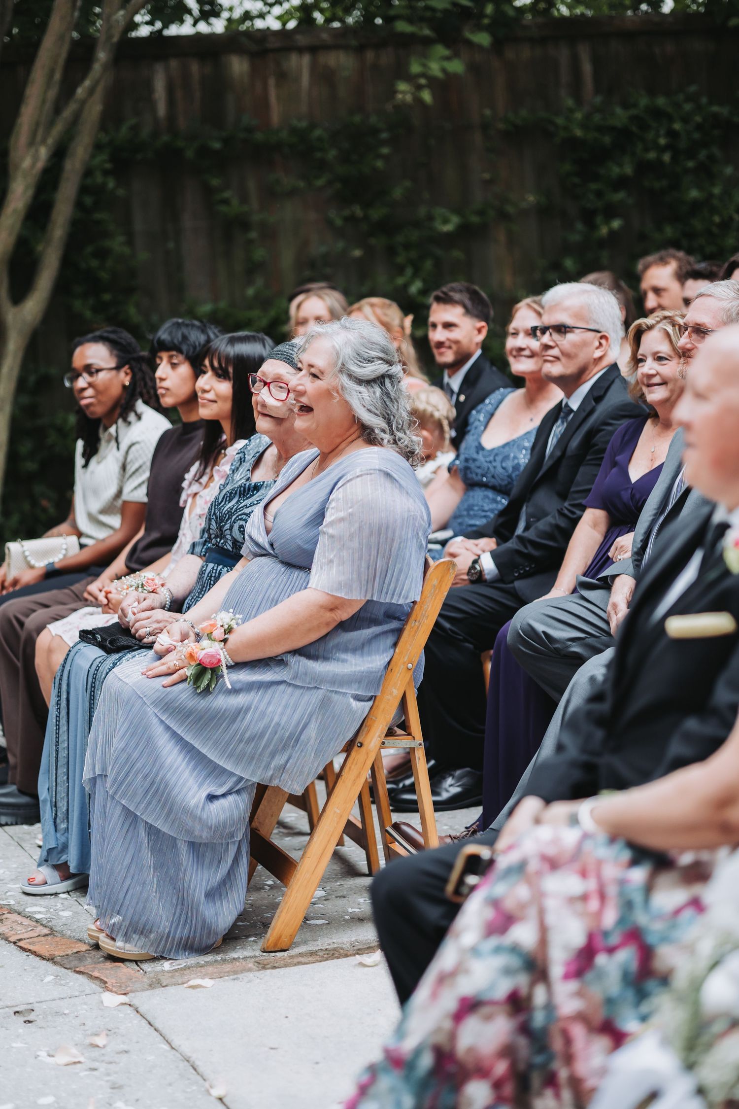 Wedding guests seated in wooden folding chairs smile during outdoor garden ceremony celebration.