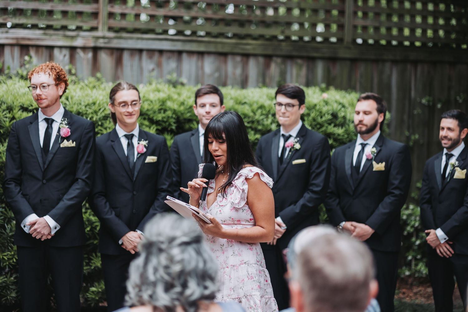 Wedding officiant reads vows as groomsmen in black suits stand behind her during an outdoor ceremony.