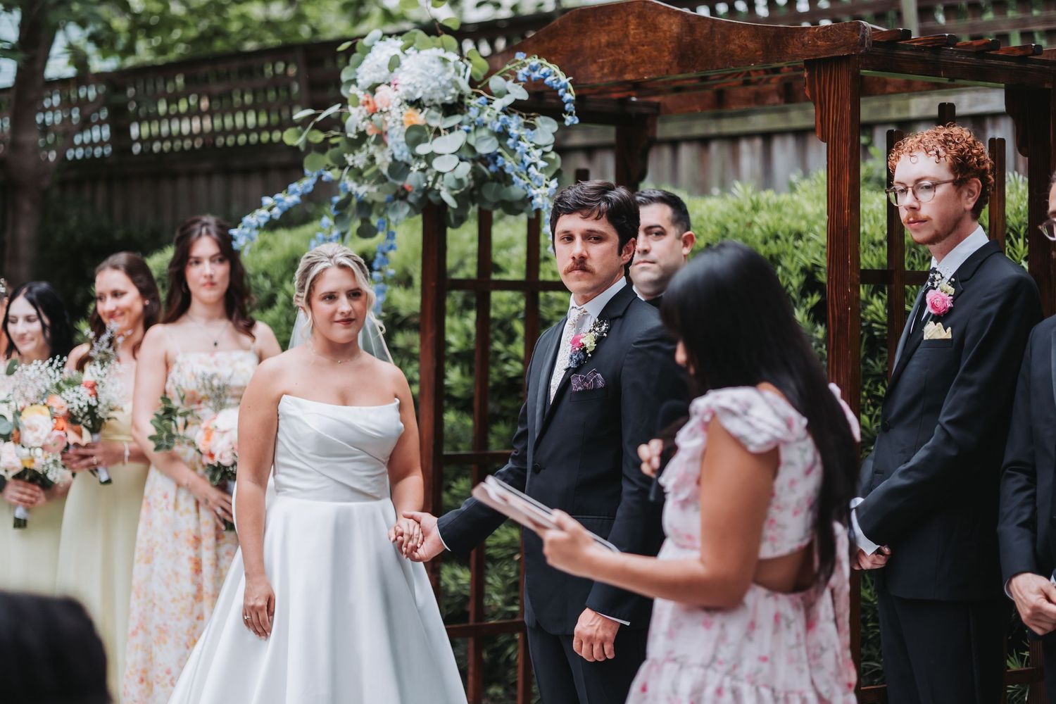 Bride and groom exchange vows at outdoor ceremony beneath blue floral arch with officiant.