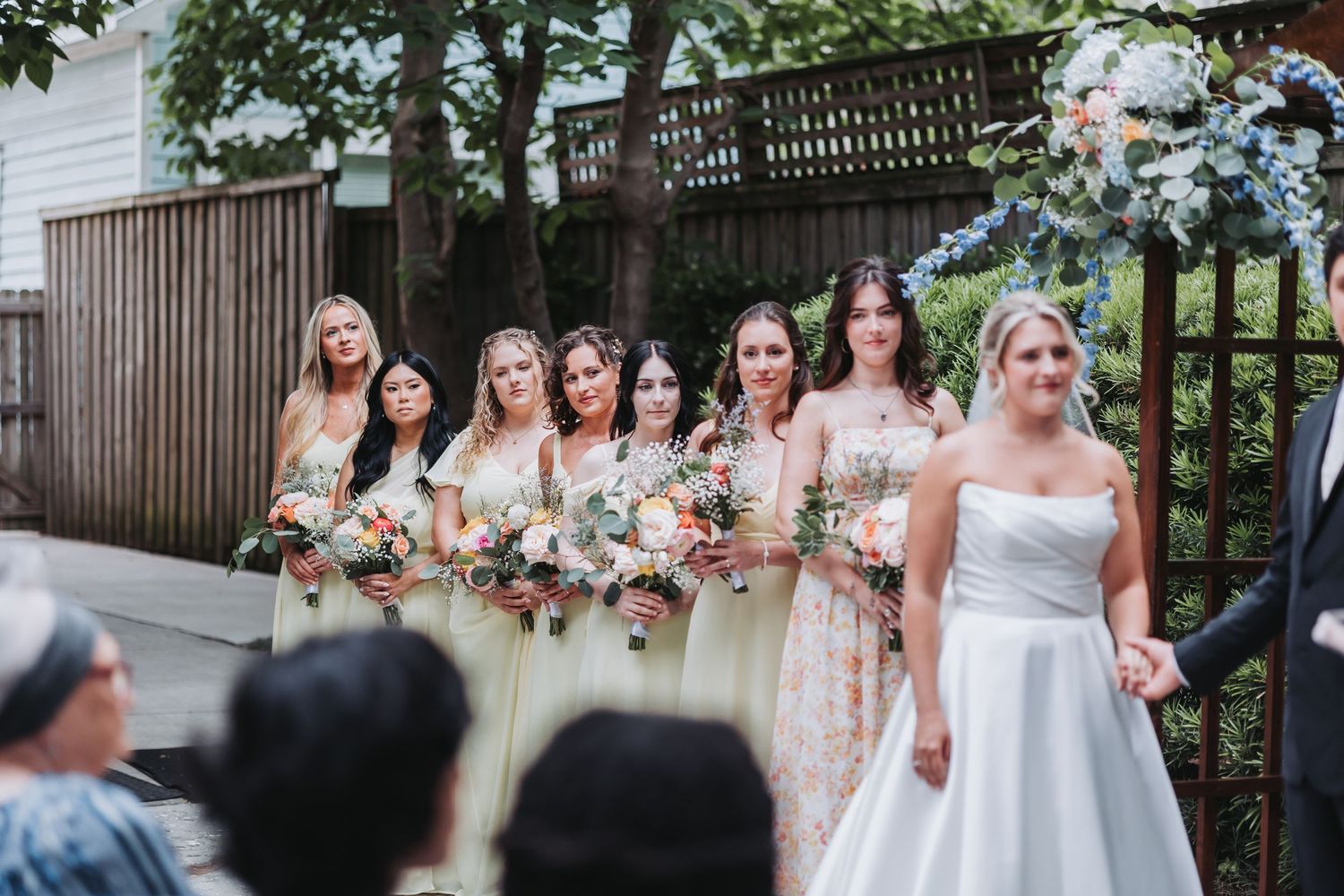 Bride in white strapless gown stands with bridesmaids in champagne dresses holding colorful bouquets.