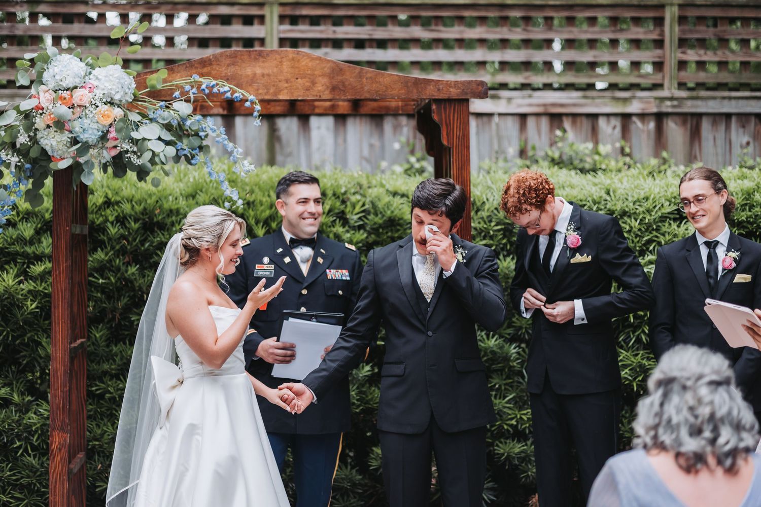 A bride and groom laugh joyfully with their wedding party at an outdoor garden ceremony.