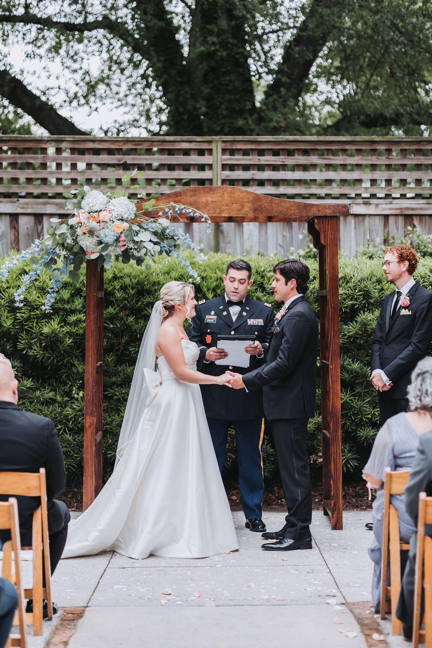 Bride and groom exchange vows outdoors under wooden arch with military officiant at garden wedding.