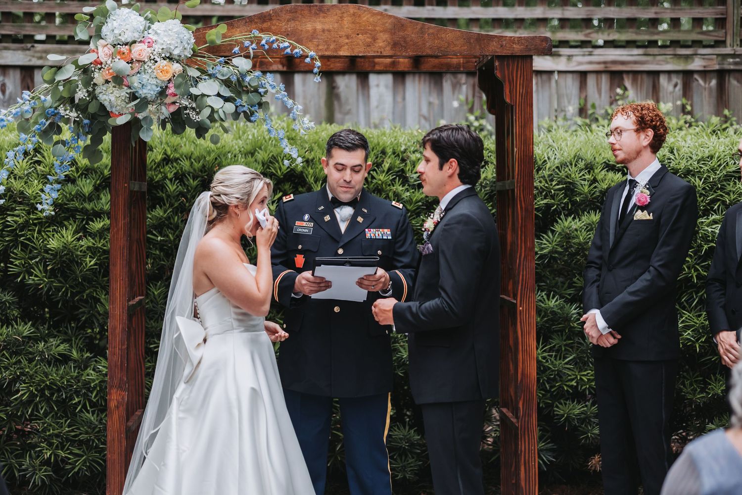 Bride and groom exchange vows under wooden arch with military officiant at outdoor wedding ceremony.