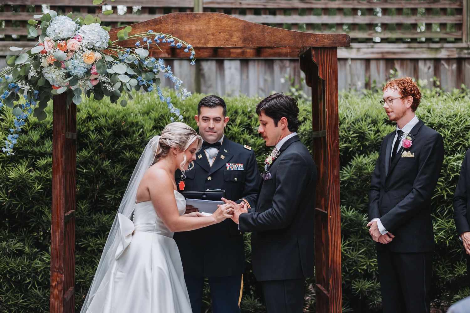 Bride and groom exchange rings at wooden arch altar during outdoor garden wedding ceremony.