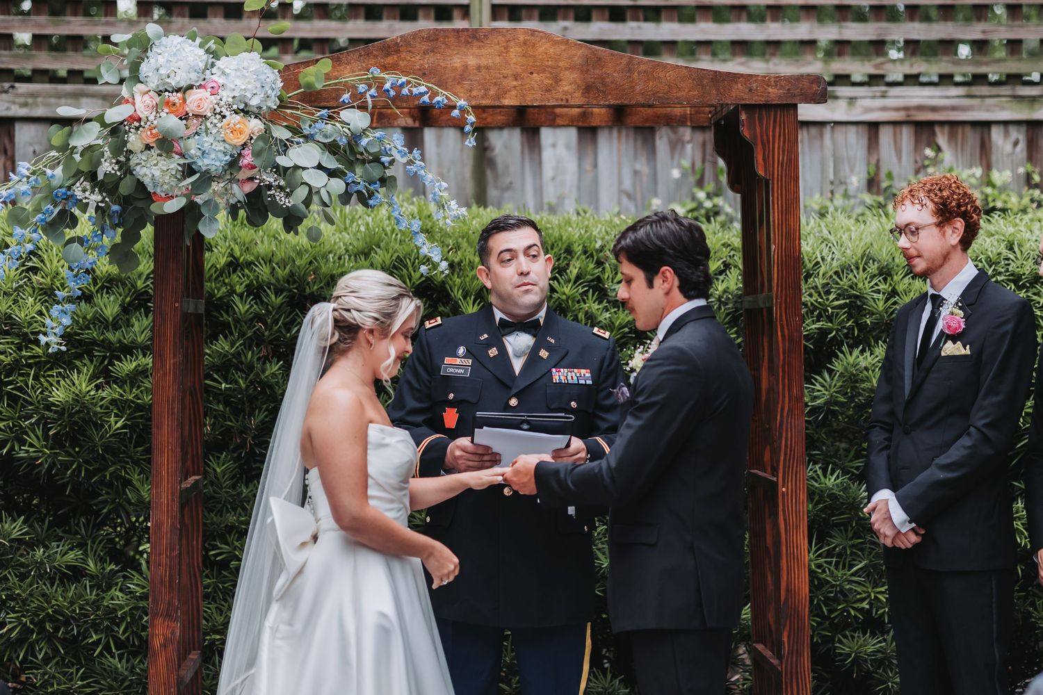 Military officiant joins bride and groom for ring exchange at rustic wooden arch outdoor wedding.