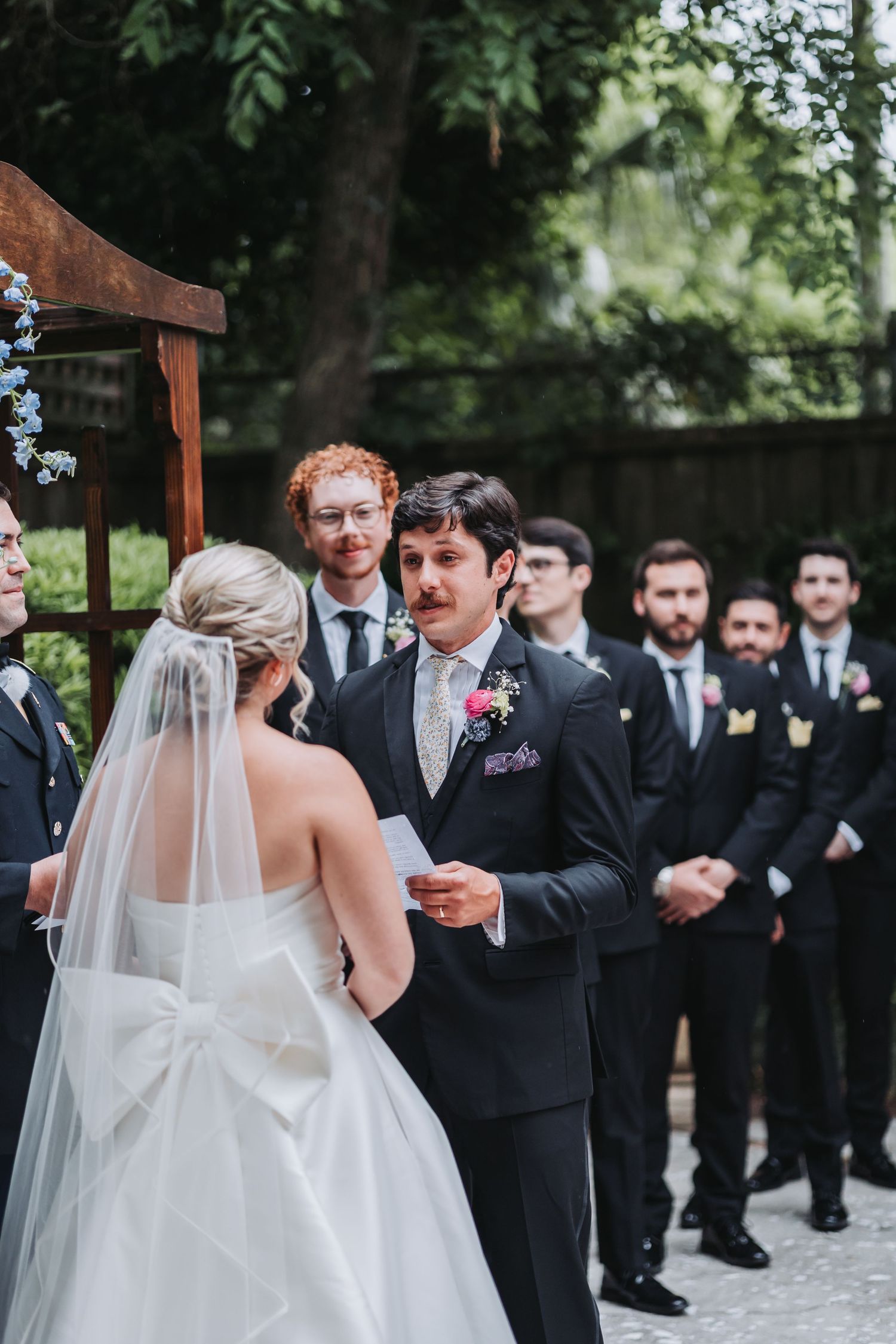 Groom reads vows to bride during outdoor wedding ceremony with groomsmen standing behind him.