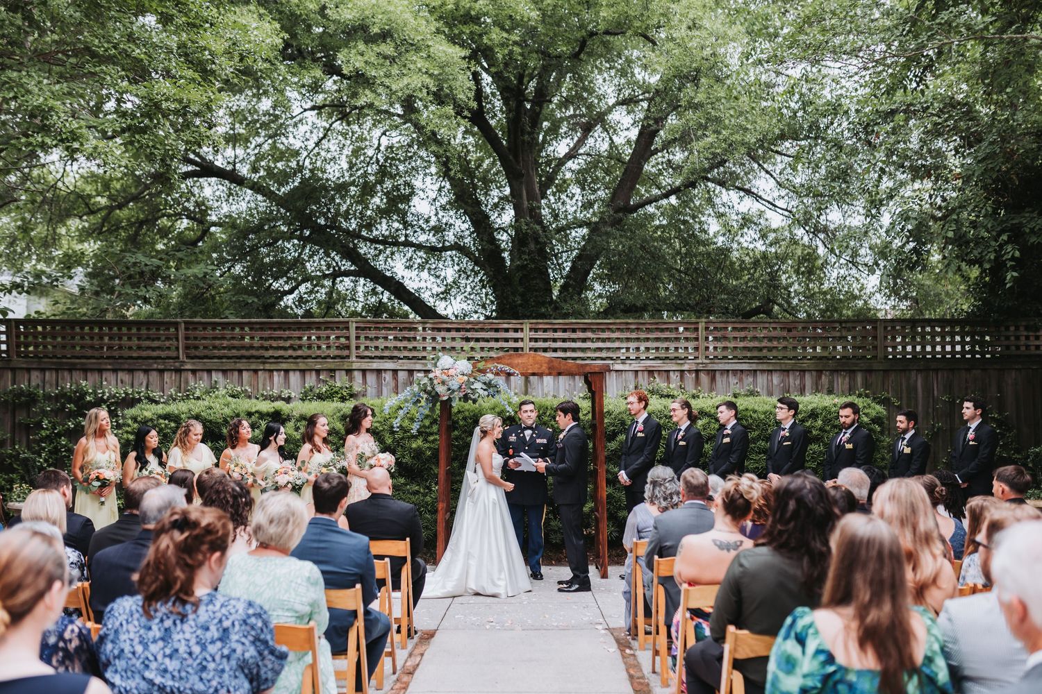 Outdoor wedding ceremony with guests seated on wooden chairs facing couple at floral arch beneath large tree.