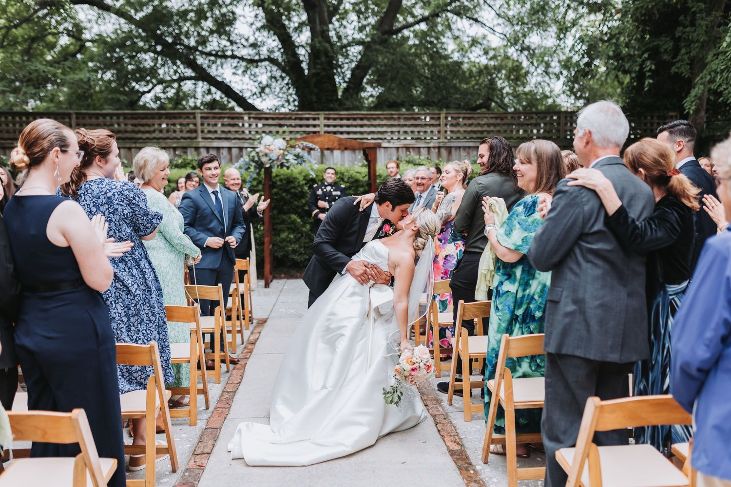 Newlyweds share romantic kiss while guests applaud along outdoor ceremony aisle after wedding.