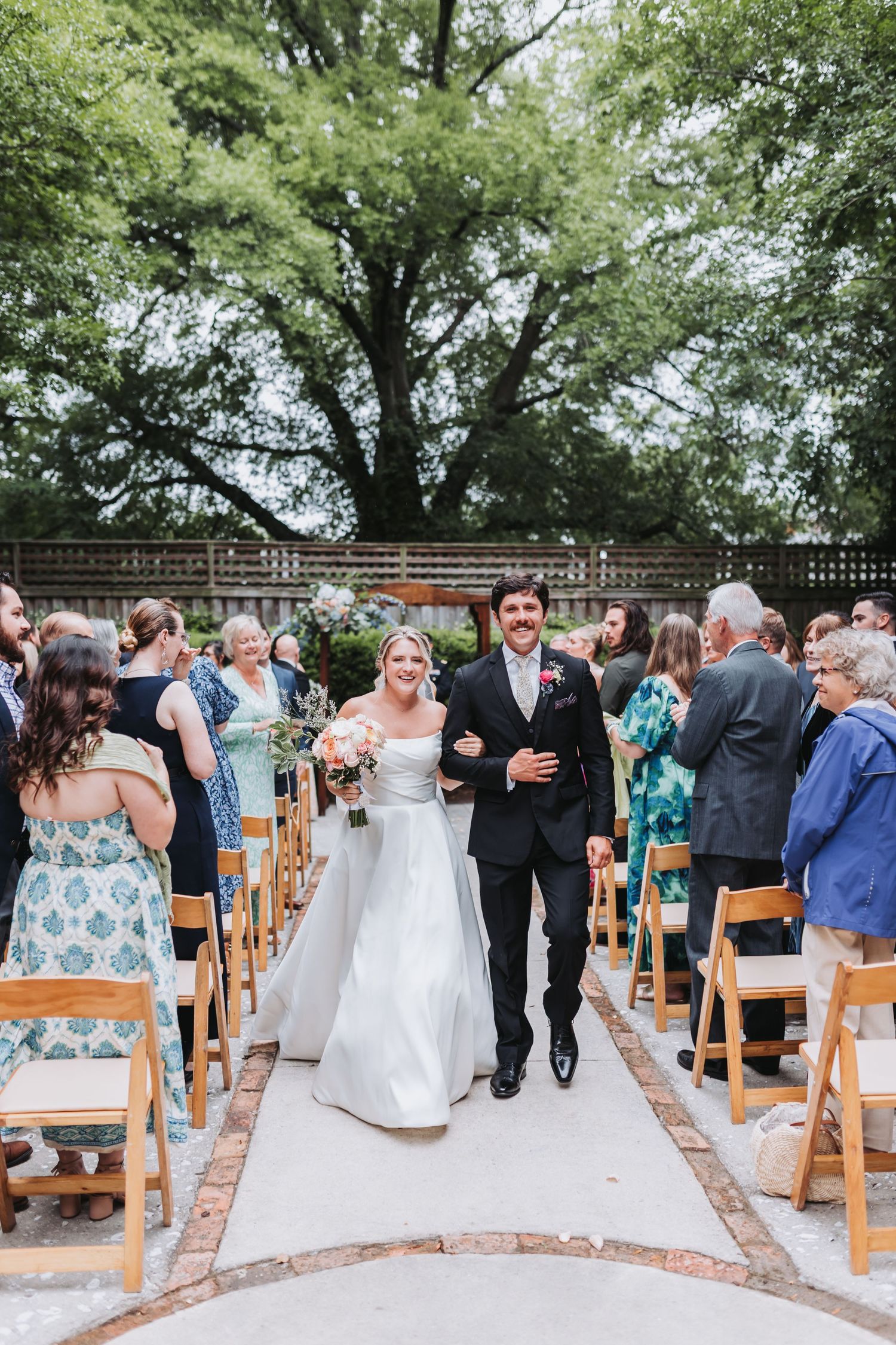 Newlywed couple walks joyfully down aisle after ceremony surrounded by standing applauding wedding guests.
