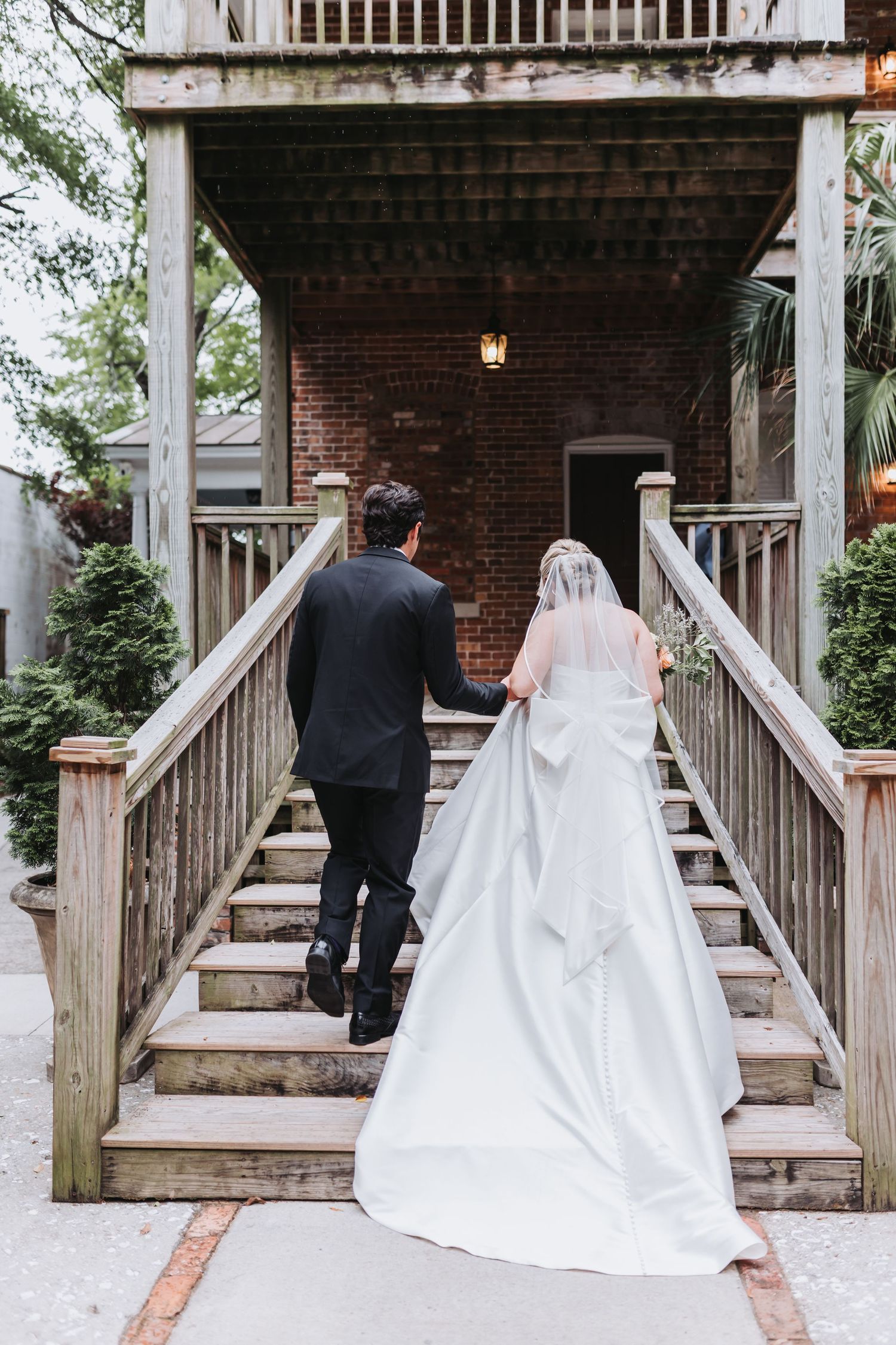 Bride and groom walk hand in hand up wooden staircase at historic brick venue after ceremony.
