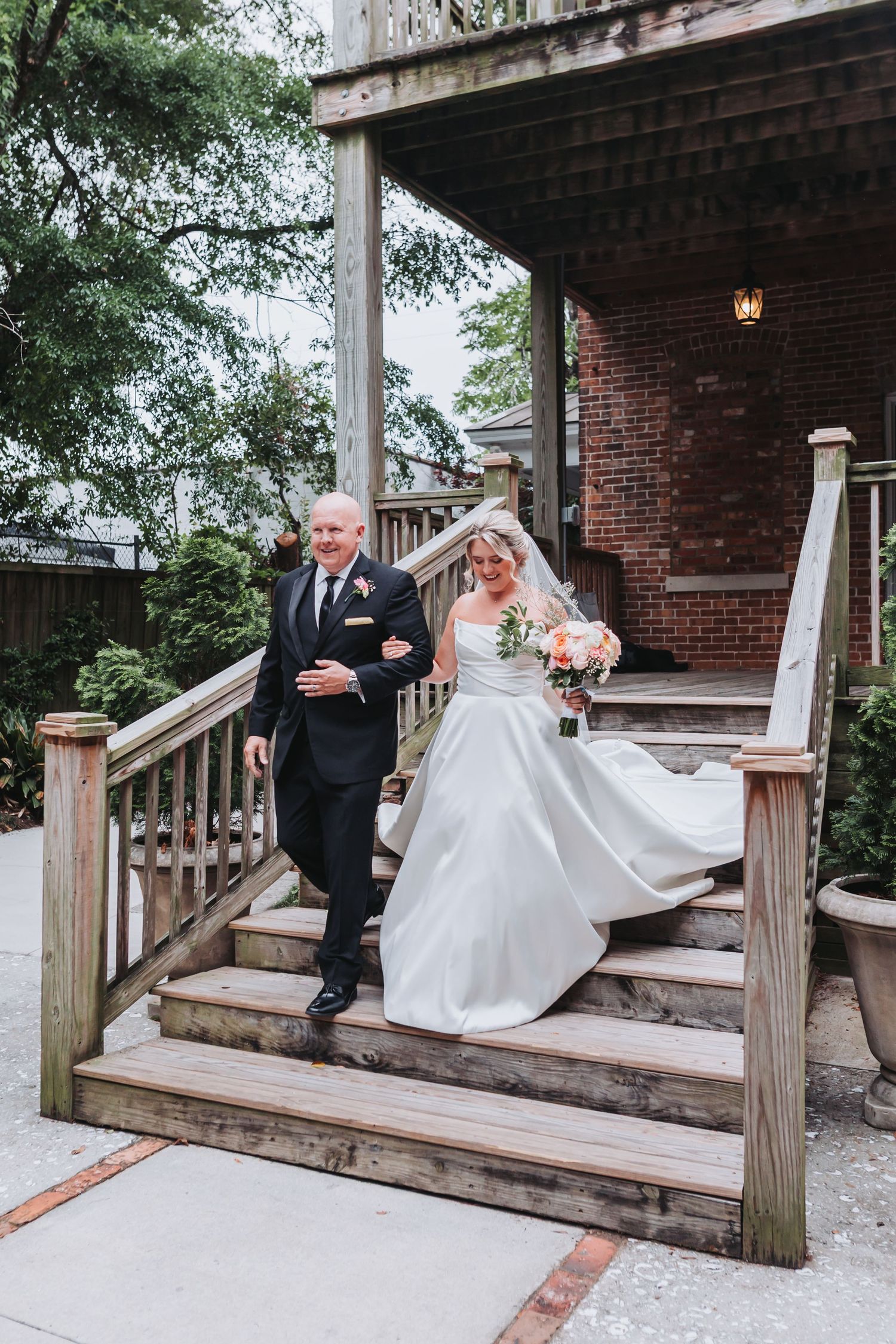 Bride in flowing white strapless gown descends wooden steps with escort at rustic brick venue.