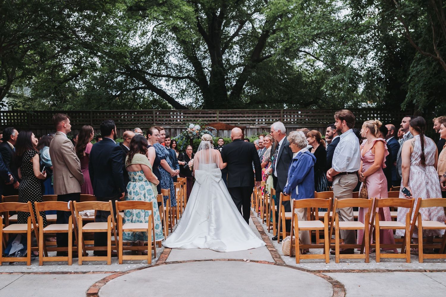 Wedding guests stand as bride walks down aisle at elegant outdoor ceremony surrounded by trees.