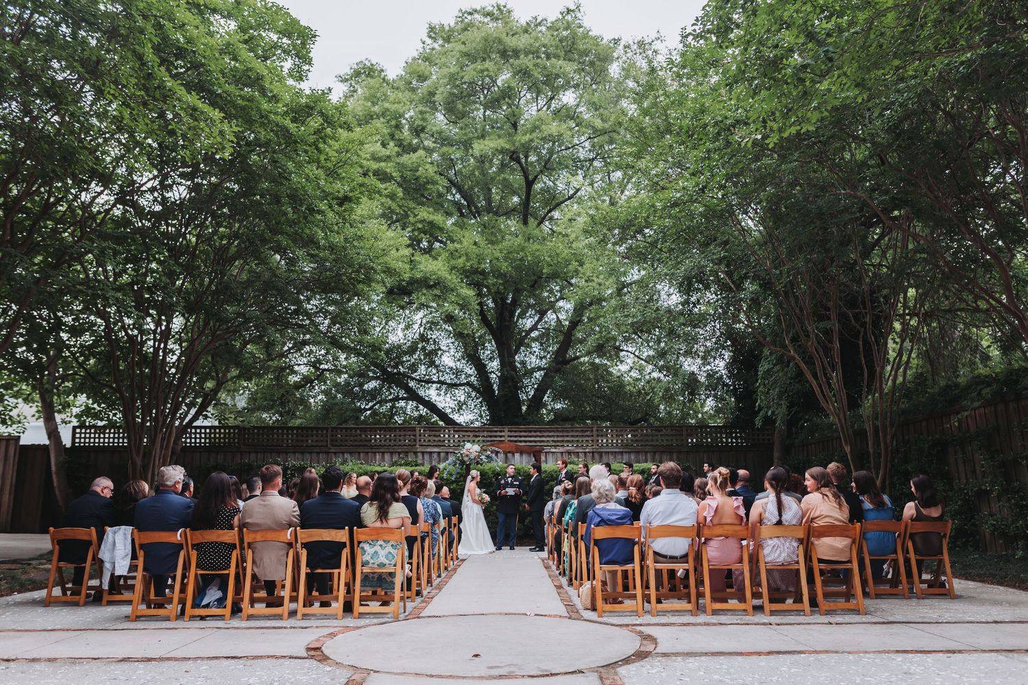 Wide shot of outdoor wedding ceremony with guests seated on wooden chairs beneath large trees.