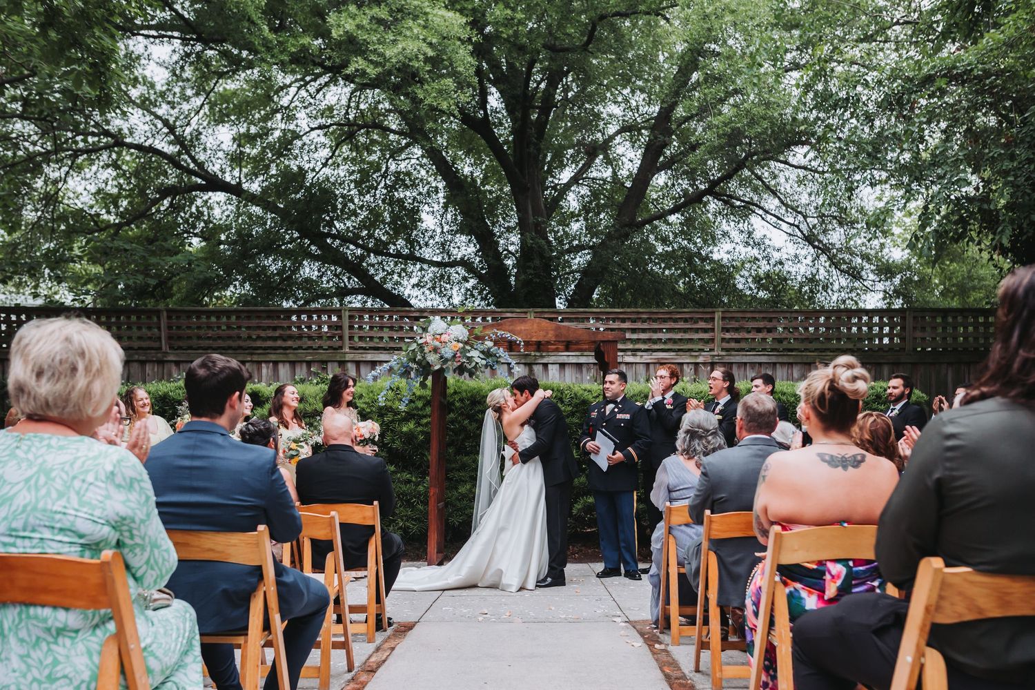 Outdoor wedding ceremony with guests seated on wooden chairs as couple exchanges vows beneath large tree.