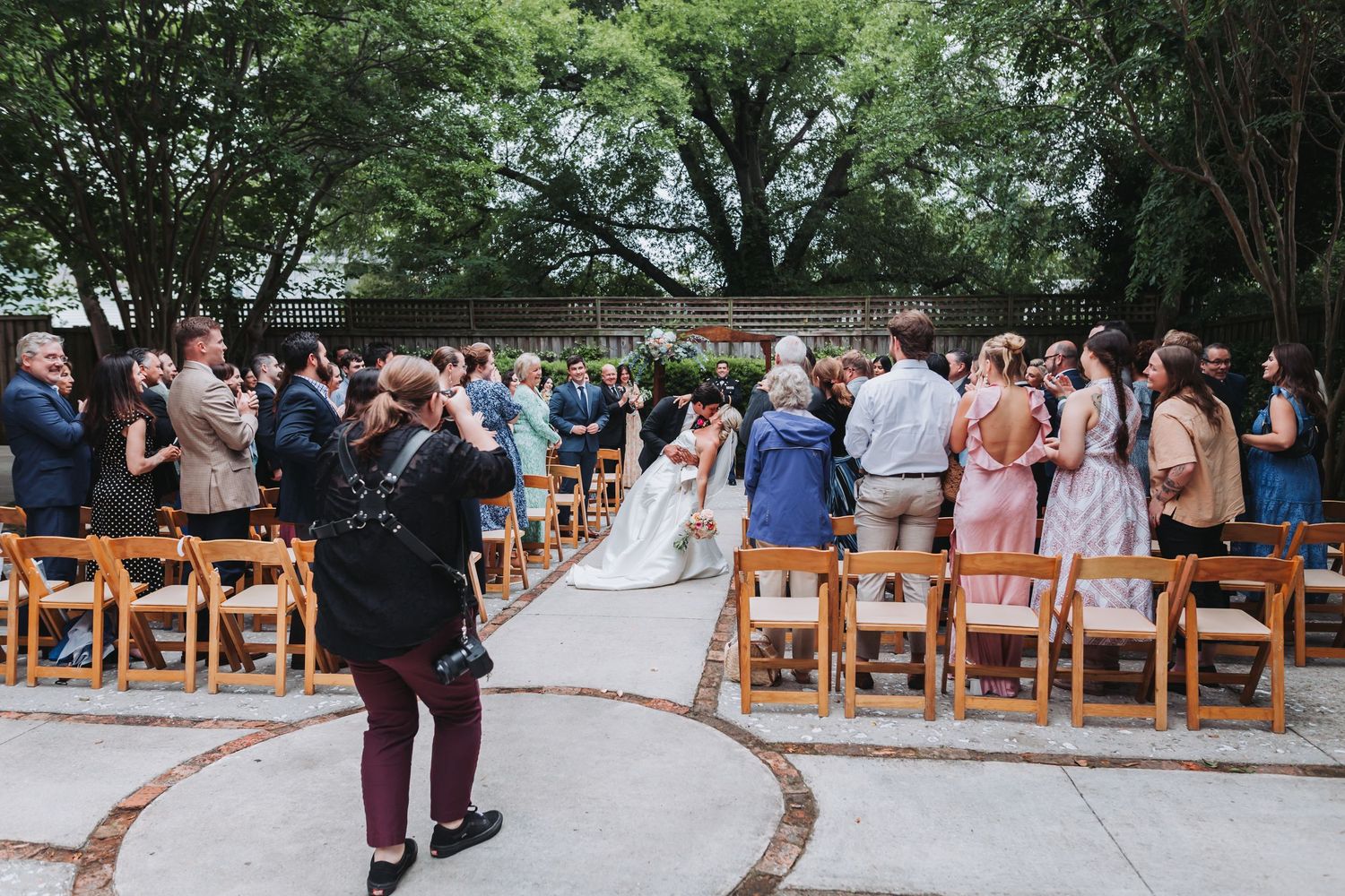 Guests stand and watch bride walk down aisle at elegant outdoor wedding ceremony with wooden chairs.