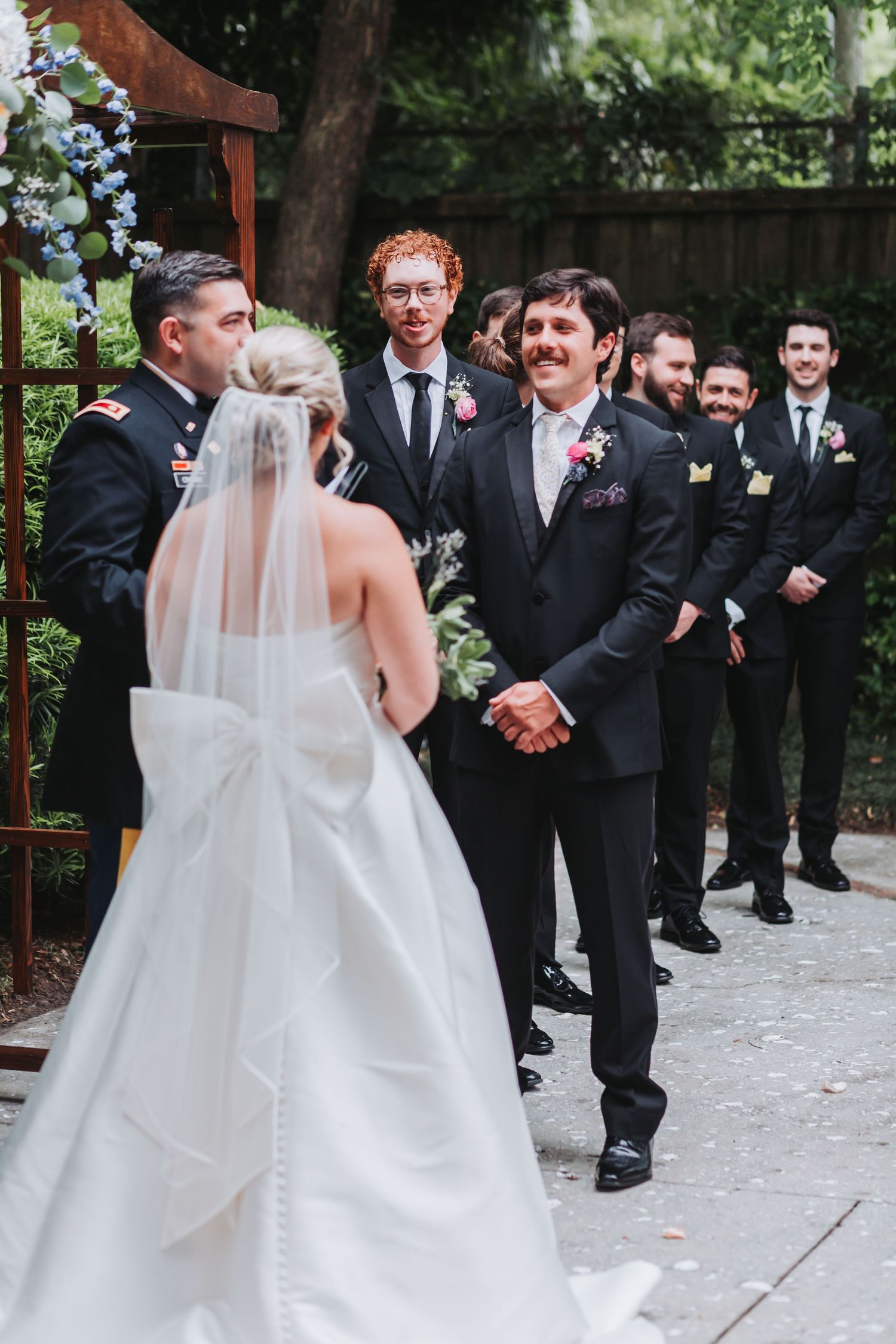 Groom smiling at bride in white gown with veil approaching altar flanked by groomsmen in black suits.
