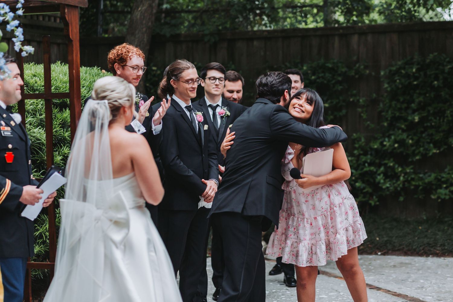 Groom embraces a guest in a floral dress while bride and wedding party look on during outdoor ceremony.