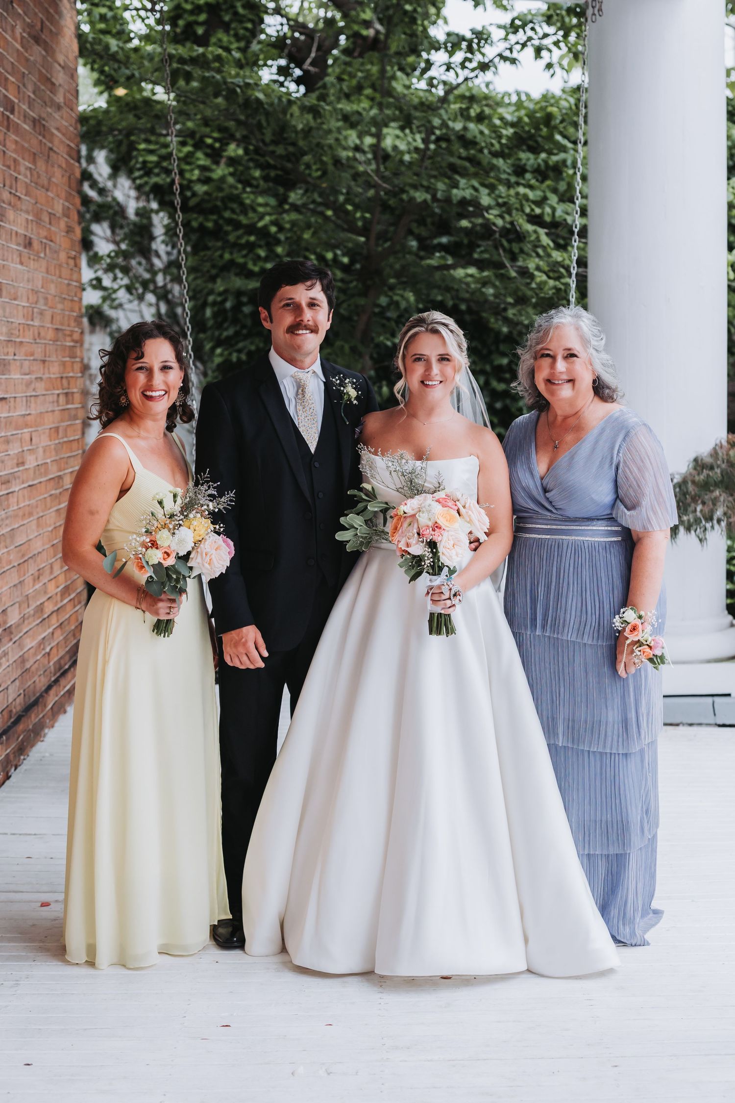 A bride and groom pose with two women in formal gowns outside a brick building with columns.