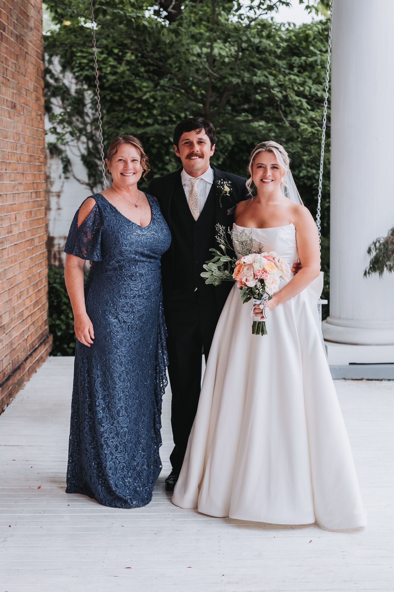 Bride in white gown holds bouquet beside groom in black suit and woman in blue lace dress at wedding.