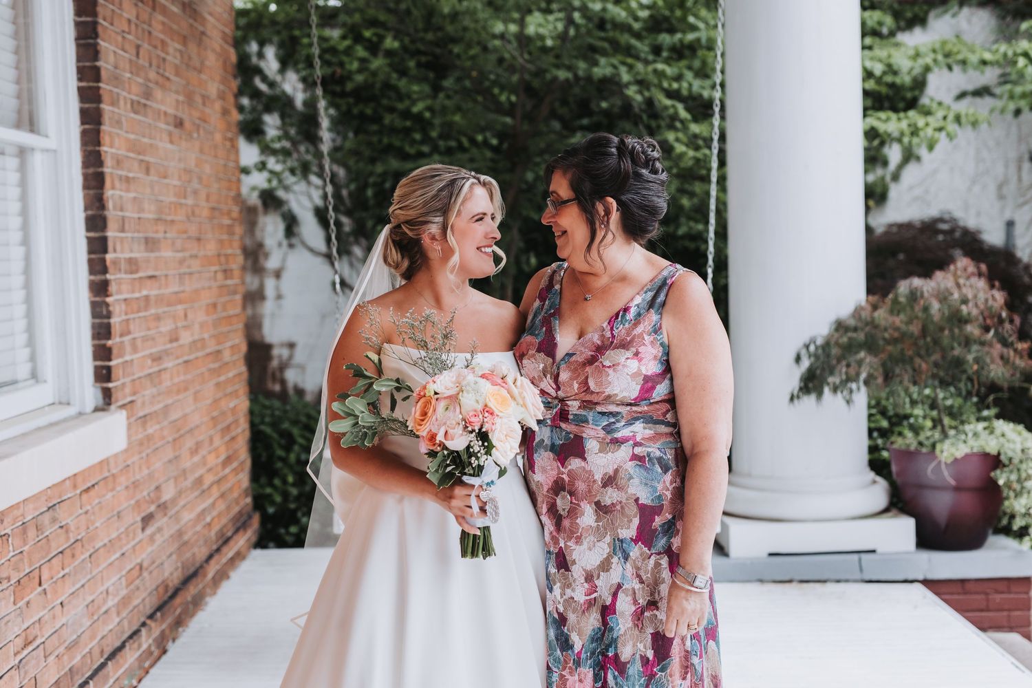 Smiling bride in strapless gown holds pink bouquet standing with woman in floral dress outdoors.