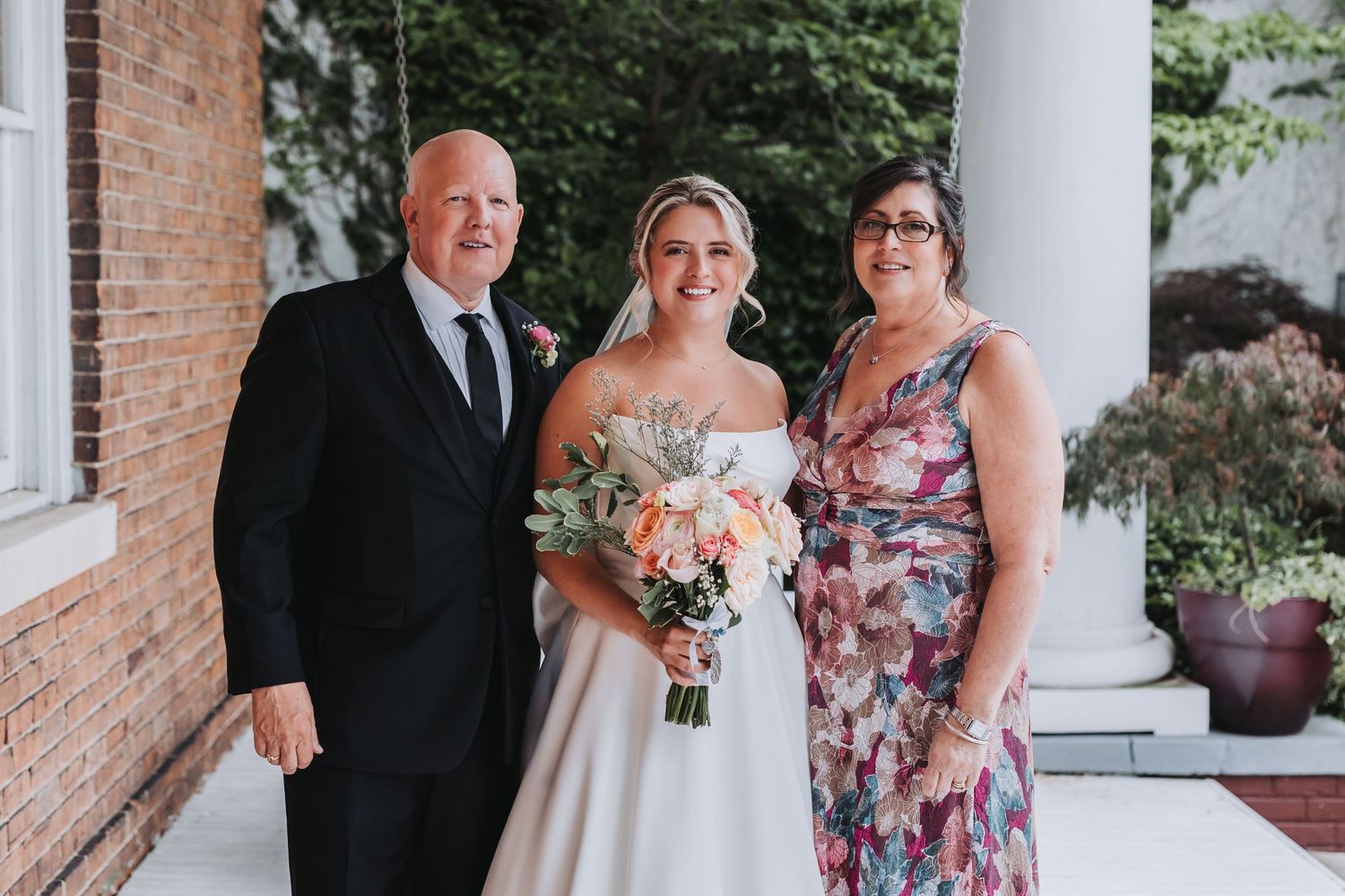 Bride poses with bouquet between two guests outside a brick building with white columns.