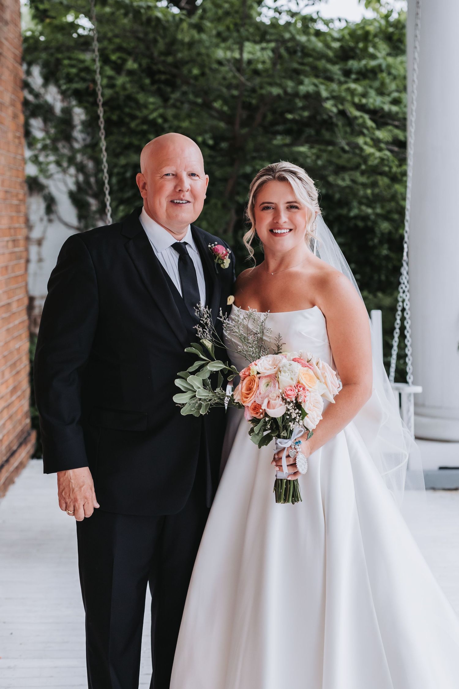 Bride in strapless white gown holds peach bouquet beside older man in black suit on white porch.