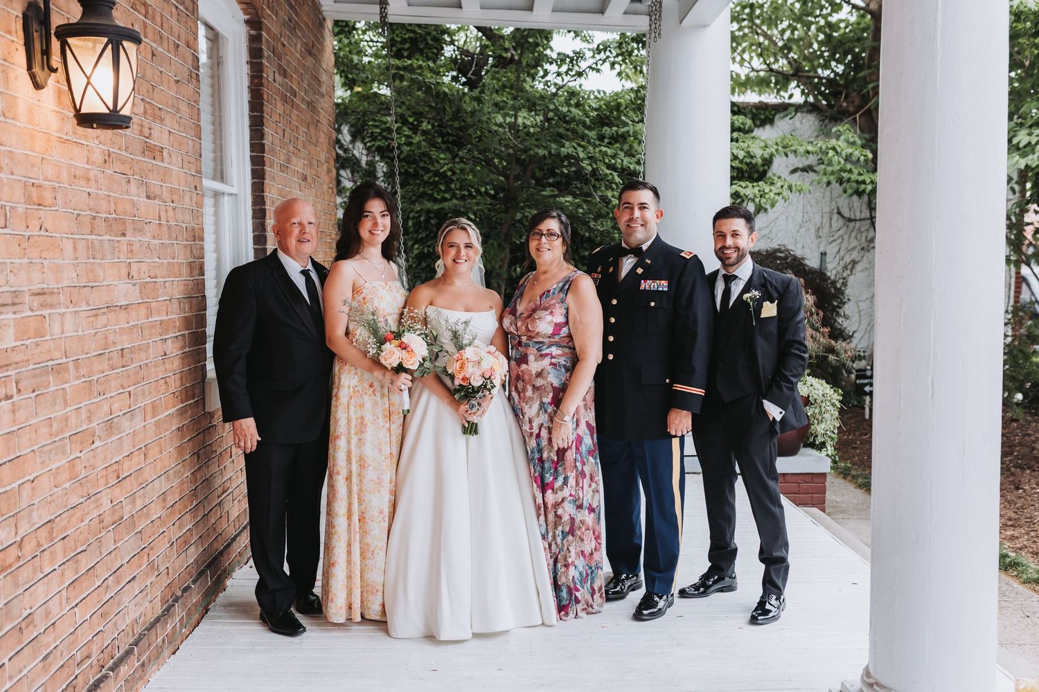 Wedding party poses together on a brick porch, including bride in white gown and groom in military dress uniform.