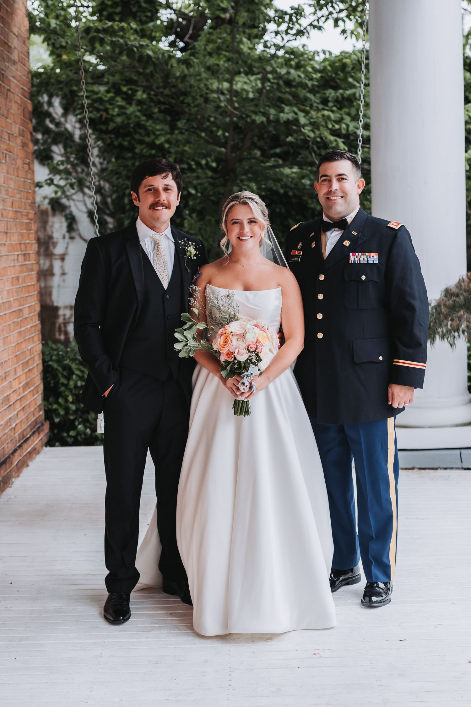 Bride in white ball gown poses with two groomsmen, one in military dress uniform, holding bouquet.