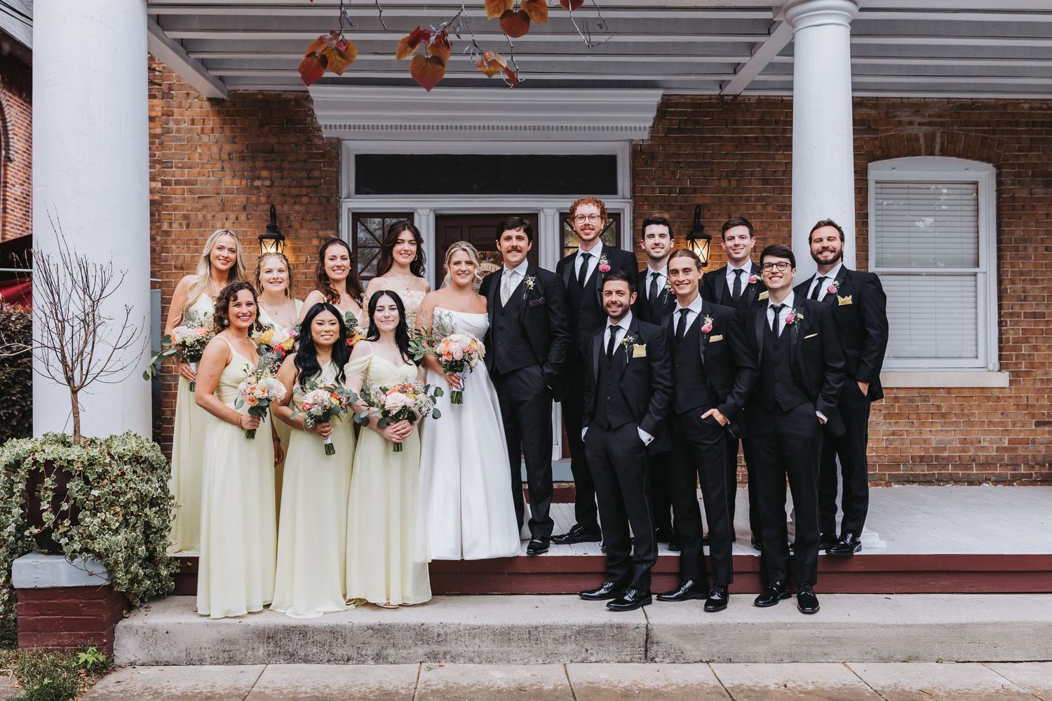 Wedding party poses on steps of brick building with bridesmaids in cream gowns and groomsmen in black.