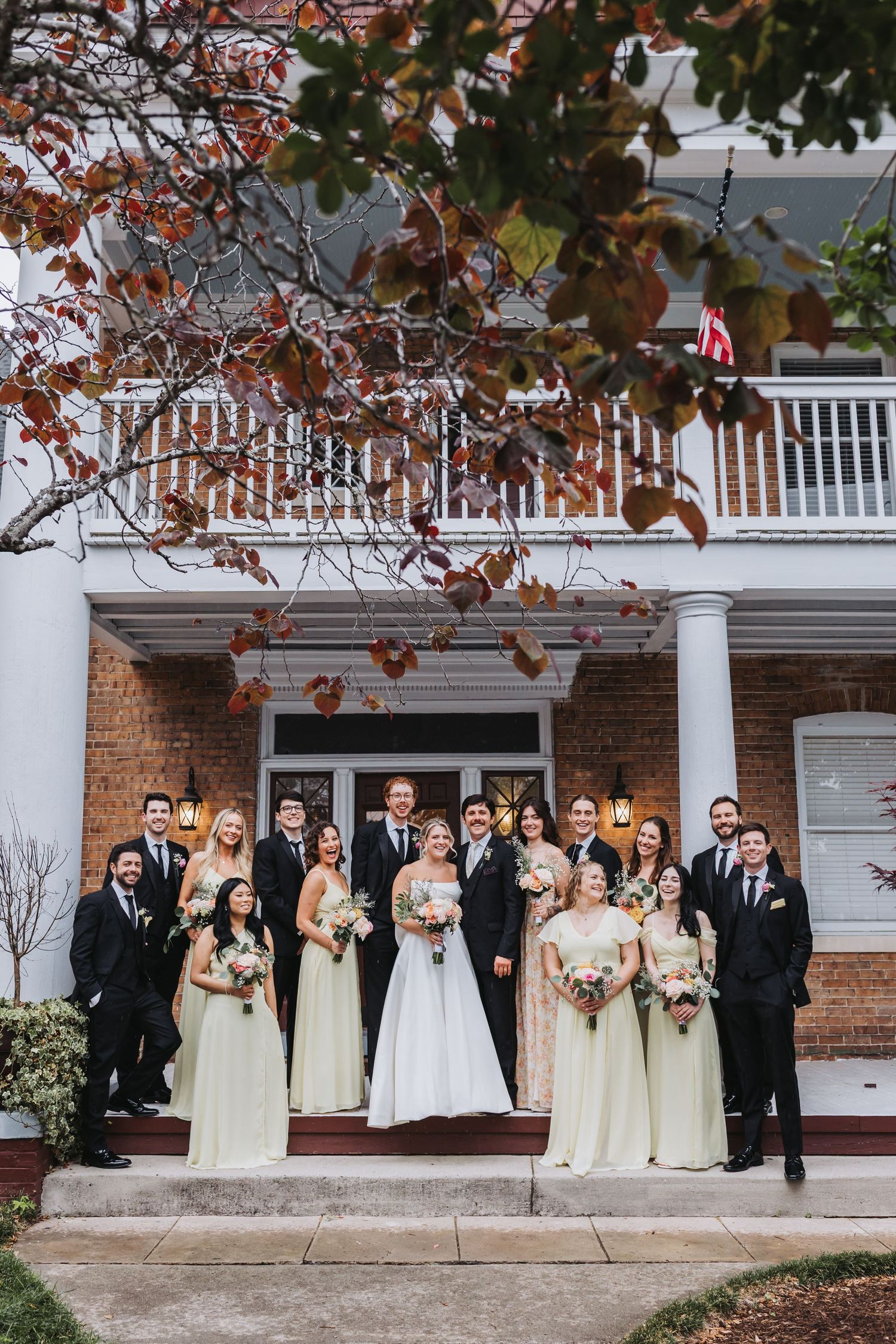 Wedding party poses in front of brick venue, bridesmaids in sage green gowns, groomsmen in black suits.