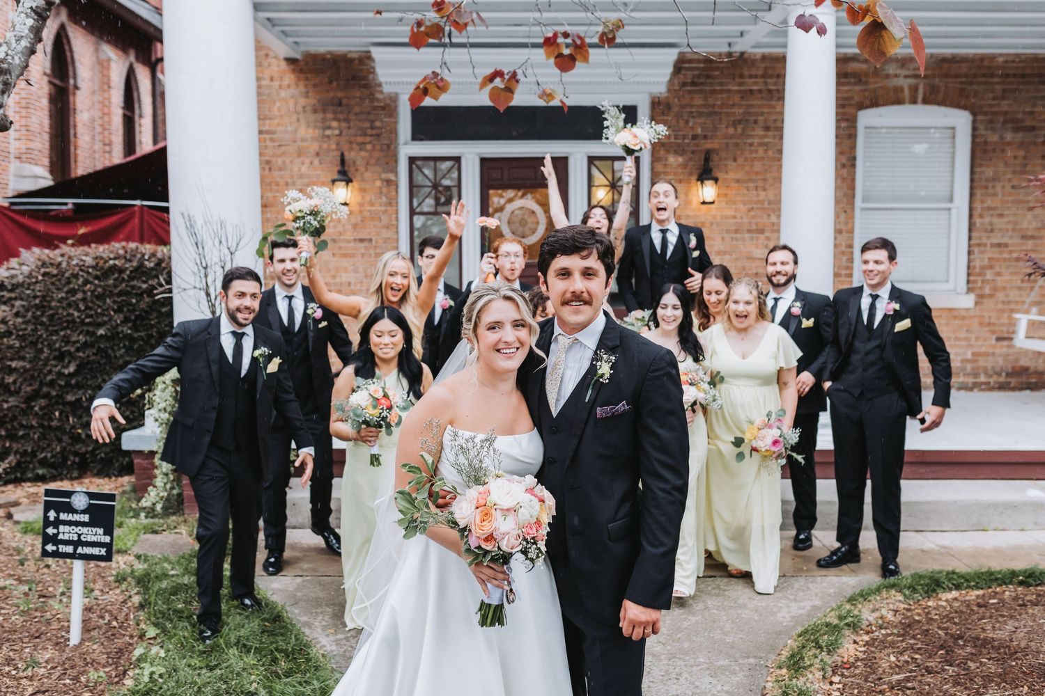 Bride and groom pose with full wedding party outside brick venue with fall floral decorations.