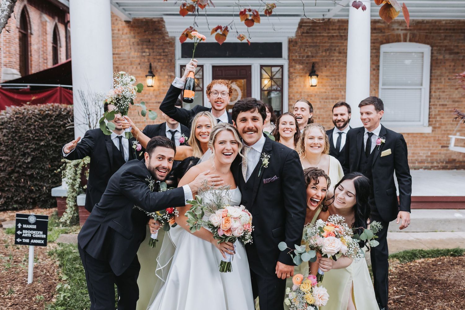 Bride and groom celebrate with wedding party outside brick venue holding champagne bottle.