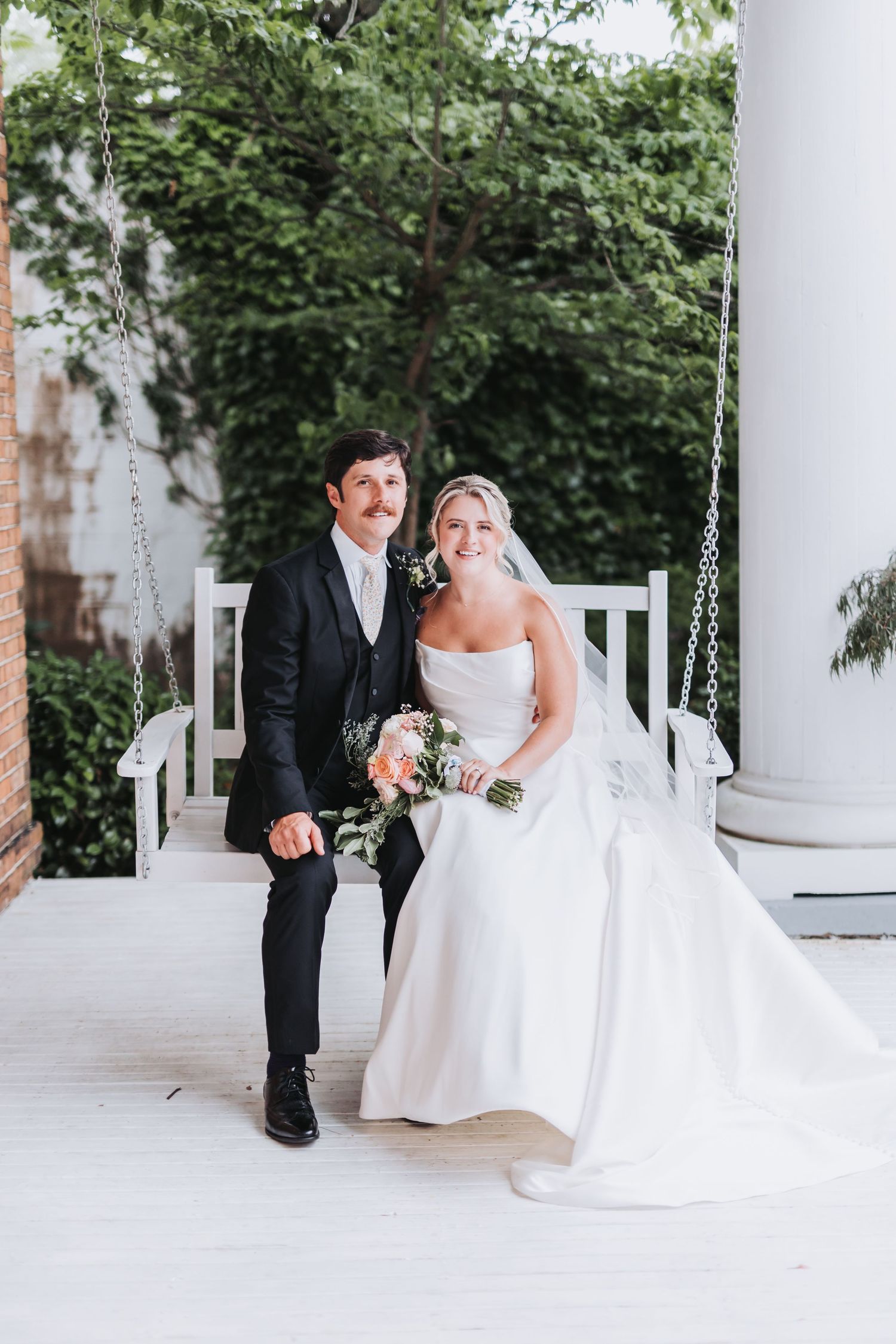 Bride and groom sit together on white porch swing surrounded by lush green foliage backdrop.
