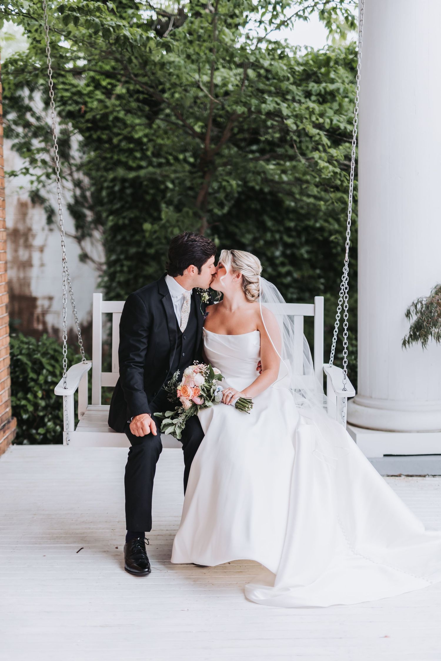 Bride and groom share a kiss on a white porch swing surrounded by lush green foliage.
