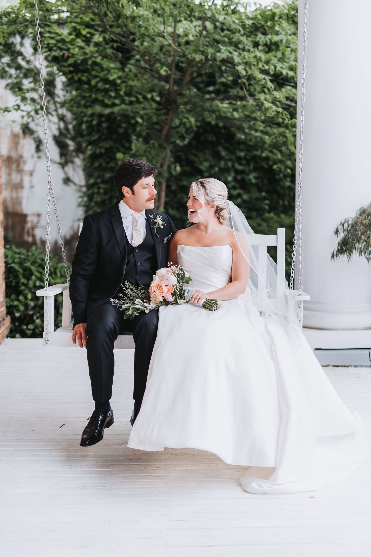 Bride and groom laugh together on a white porch swing on their wedding day.