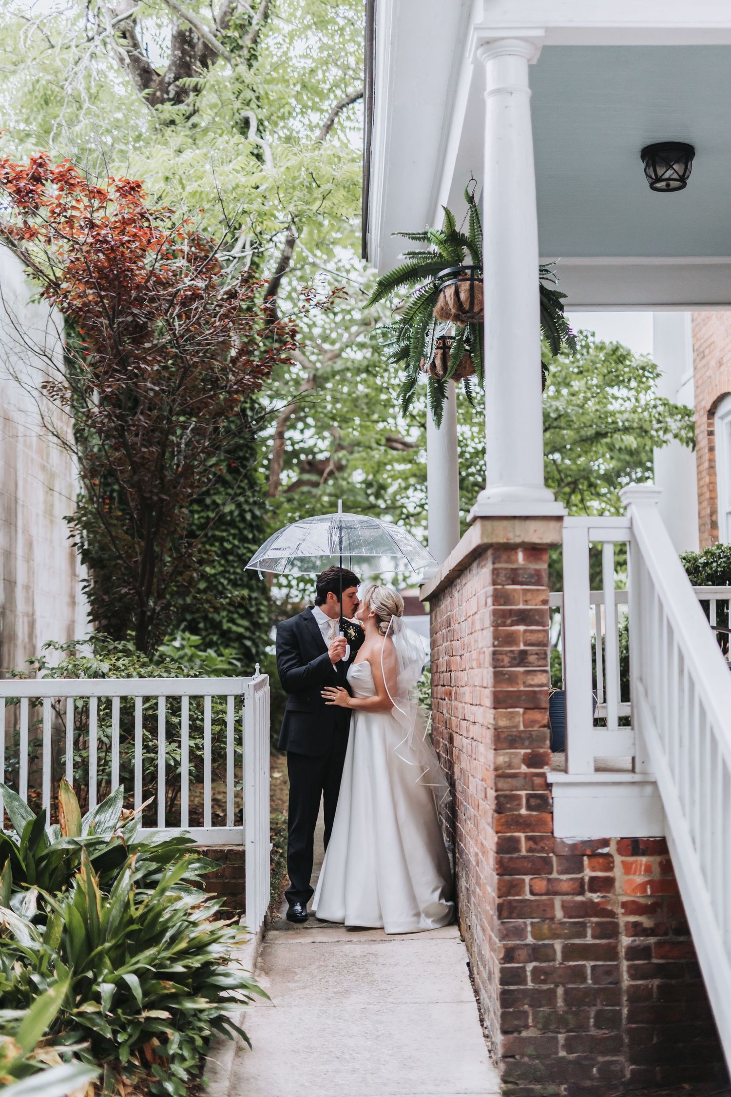 Bride and groom share romantic kiss under clear umbrella on garden porch amid lush greenery.