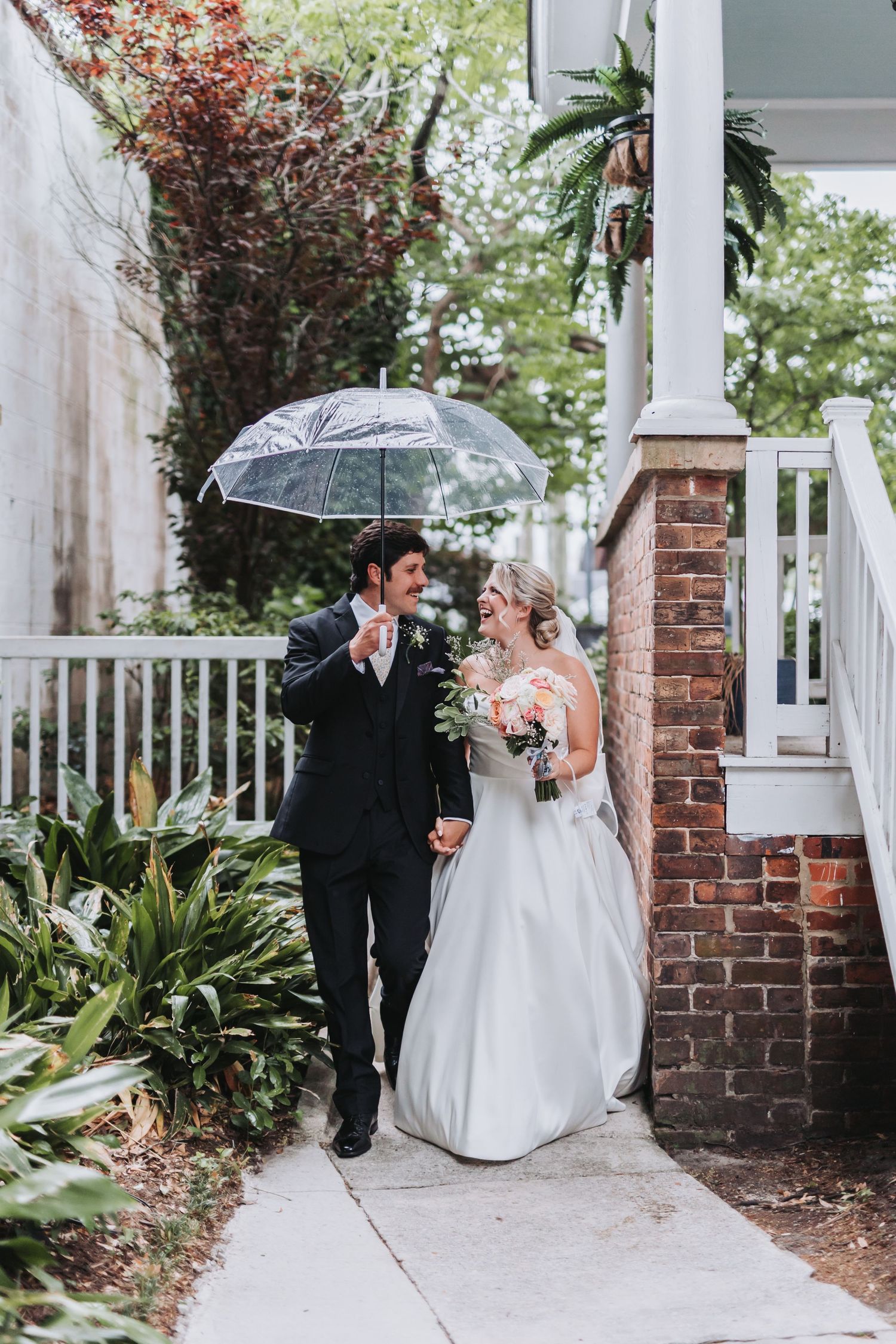 Bride and groom share a laugh walking under a clear umbrella on their rainy wedding day.