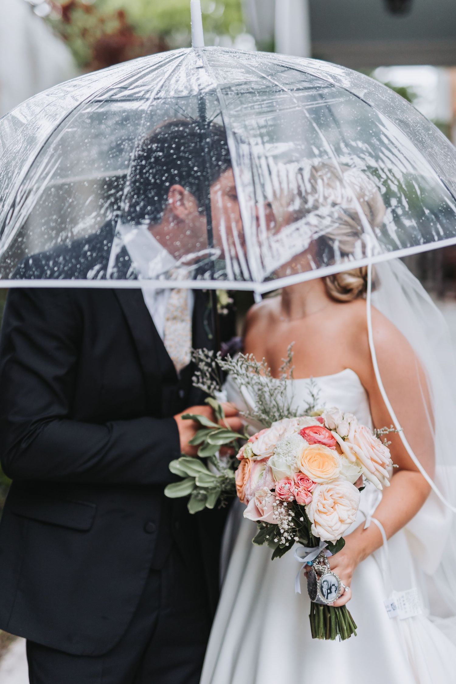 Bride and groom sharing a kiss under a clear umbrella on their rainy wedding day.