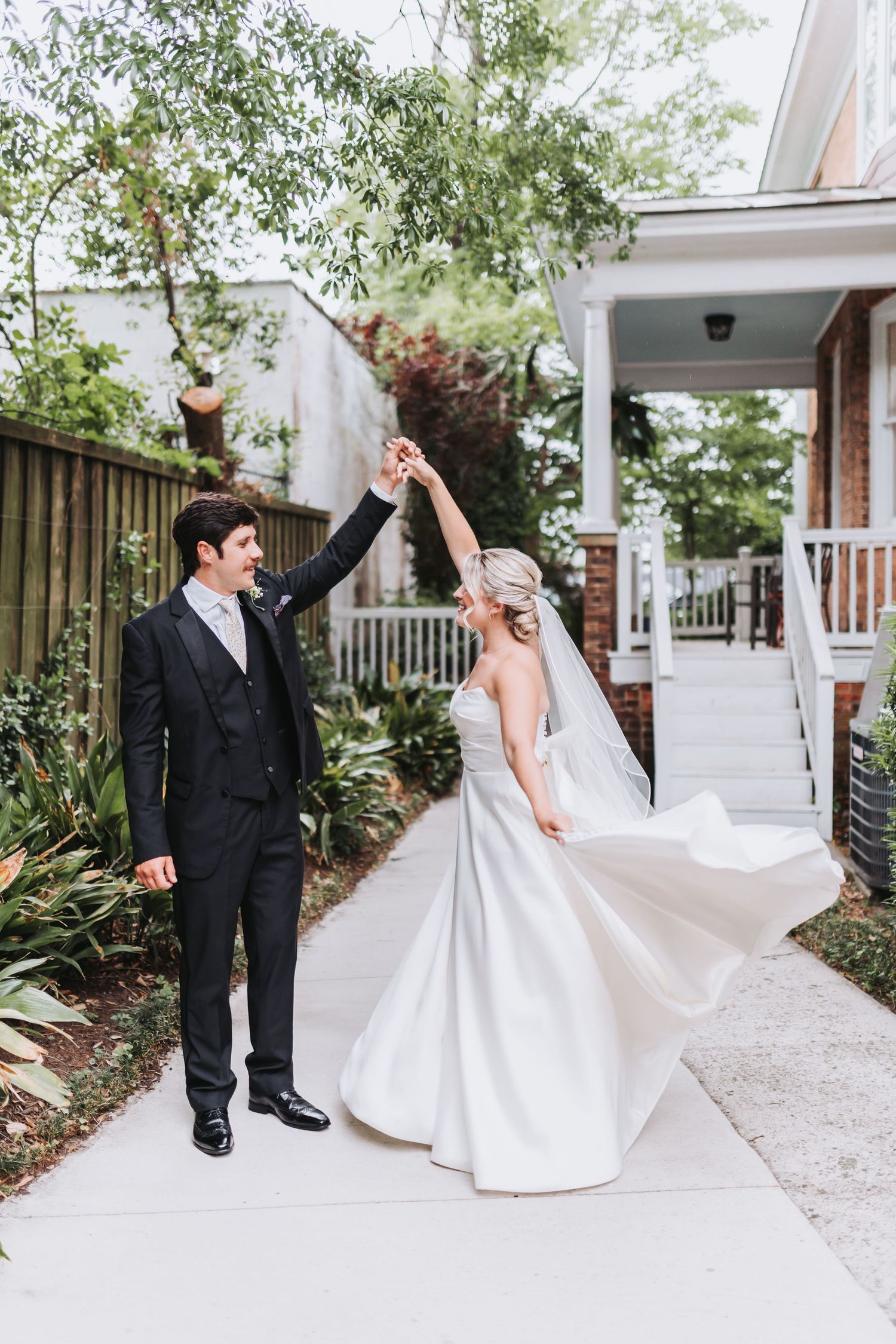 A groom in a black suit twirls his bride in a white gown and veil in a lush outdoor garden setting.